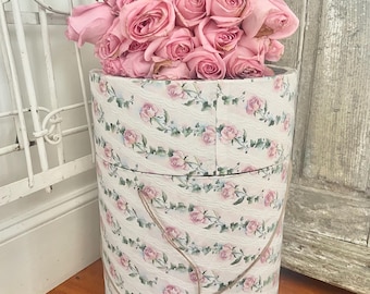May include: A tall, cylindrical hat box overflowing with pink roses. The box is decorated with a white floral pattern featuring pink roses and has a twine handle. The roses are a vibrant pink. The box sits on a wooden floor.