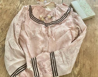 May include: A vintage pink blouse with long sleeves and a ruffled collar. The collar and cuffs feature three dark brown stripes. The blouse is adorned with dried flowers and rests on a wooden surface.