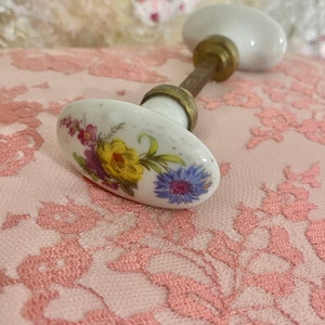 May include: White porcelain door knob with a floral design featuring yellow, purple, and blue flowers. The knob has a brass stem.