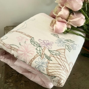 May include: Folded white and pink floral embroidered quilt with a basket design. The quilt features pink, blue, and purple flowers with green leaves. A bouquet of pink roses rests on top of the quilt.