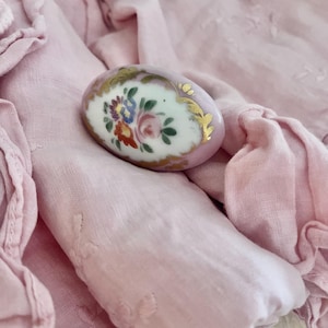 Może przedstawiać: Owalna, różowa broszka porcelanowa z ręcznie malowanym kwiatowym wzorem. Broszka ma biały środek z kolorowymi kwiatami i złotymi akcentami. Leży na miękkiej, różowej tkaninie.