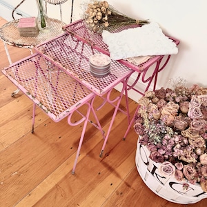 May include: A set of vintage pink metal side tables with a diamond mesh top. The tables are next to a white wire chair and a basket of dried flowers. A vase of pink roses and a small tin are also visible.