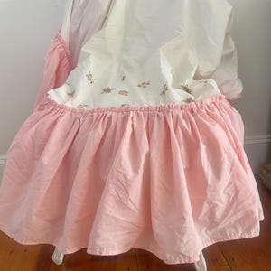May include: A pink and white crib skirt with a floral pattern. The skirt has a ruffled edge and is draped over a white crib. The fabric appears to be cotton or a similar material.