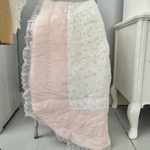 May include: A pink and white quilted baby blanket with a floral pattern and lace trim. The blanket is draped over a small, antique chair. The pink section features quilted floral designs and a scalloped edge.