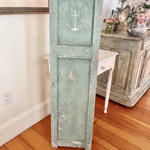 Puede incluir: Un panel de puerta de madera verde agua desgastado con un recorte de ancla blanco. La pintura se está descascarillando, revelando la madera debajo. El panel está de pie sobre un suelo de madera.