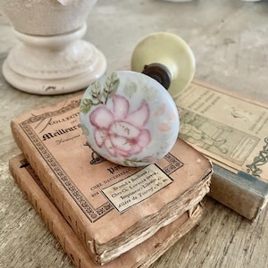 以下が含まれることがあります： 手描きのピンクの花柄が施された、ヴィンテージの磁器製ドアノブ。古い本の上に置かれています。ノブはクリーム色のベースと金属製のネジが付いています。本は古びた茶色の表紙で、フランス語のテキストが書かれています。