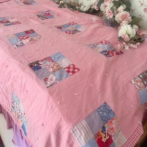 Op de afbeelding: Een roze patchworkquilt met verschillende stoffen vierkanten in bloemen- en patroonontwerpen. De quilt heeft een effen roze achtergrond met decoratieve stiksels en een ruitjesrand. Het bed is bedekt met een roze sprei.