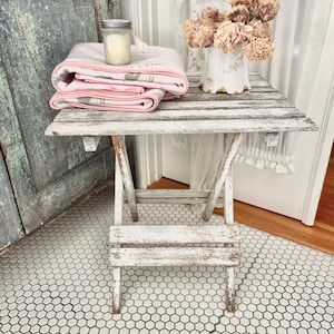 Könnte beinhalten: Ein kleiner, weißer Holztisch mit Gebrauchsspuren, mit einer gefalteten rosa Decke, einem Glas mit Deckel und einer Vase mit getrockneten Blumen. Der Tisch steht auf einem weiß-schwarzen Sechseck-Fliesenboden.