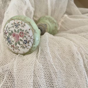 May include: A round porcelain door knob featuring a pale green border and a central white circular panel decorated with a hand-painted floral motif in shades of pink, red, blue, and green, with a metallic spindle extending from the back.