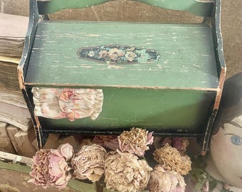 May include: A distressed green wooden box with a handle, featuring a floral design on the lid. The box is filled with dried flowers and sits atop a painted wooden container. The overall aesthetic is vintage and rustic.