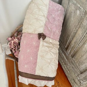 Op de afbeelding: Een opgevouwen, gewatteerde babydeken met een crème- en roze bloemenpatroon. De deken heeft een bruine strik en bies, met een witte gefranjerde rand. De deken is opgevouwen en leunt tegen een muur.