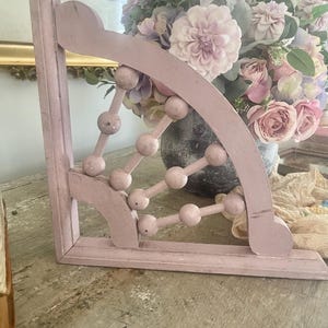 Peut inclure: Équerre d'étagère d'angle rose pâle. Le support présente un design décoratif avec des éléments arrondis et un motif géométrique. Le style est vintage, avec une finition vieillie. Le support est en bois.