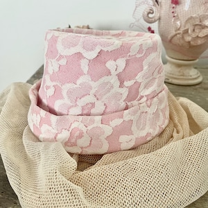 May include: A pink and white lace hat with a wide brim sits atop a beige mesh fabric. The hat features a floral lace pattern. A decorative vase is visible in the background.