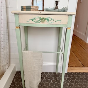May include: A tall, slender side table painted in light green and cream, featuring a single drawer with a painted green ribbon motif and a floral knob. The table has four straight legs with gold-painted accents and a lower support bar holding a white cloth.