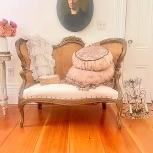 May include: An antique loveseat with a gold-toned wooden frame and cream-colored upholstery. The loveseat is adorned with pink pillows and a white, ruffled textile. A vintage portrait hangs above the loveseat.