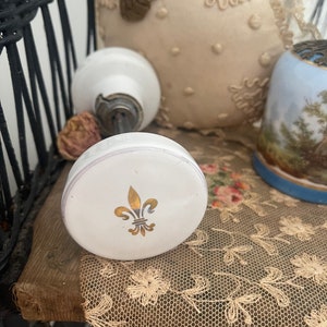 Peut inclure: Bouton de porte en porcelaine blanche avec un motif de fleur de lys dorée. Le bouton est posé sur une dentelle blanche.