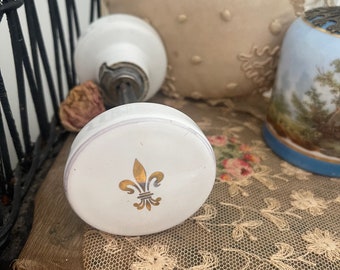 May include: White porcelain door knob with a gold fleur-de-lis design. The knob is sitting on a white lace doily.