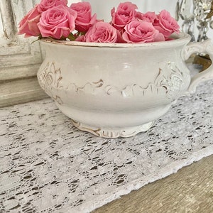Puede incluir: Un cuenco de cerámica blanco de estilo antiguo lleno de rosas rosas. El cuenco tiene un asa decorativa y patrones florales en relieve. Se encuentra sobre un camino de mesa de encaje blanco, creando una estética vintage.