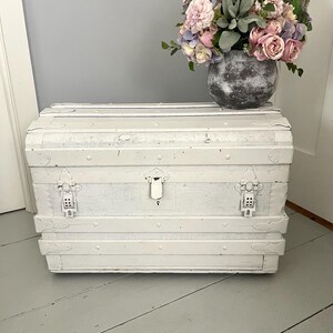May include: A white vintage trunk with metal hardware and a curved top. The trunk is painted white and has metal accents. A vase with pink, purple, and green flowers sits on top of the trunk. The trunk is on a gray wooden floor.