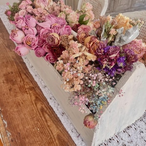 May include: A white, distressed wooden box overflowing with dried flowers in shades of pink, purple, and peach. The arrangement includes roses, baby's breath, and other dried blooms. The box sits on a white lace runner over a wooden table.