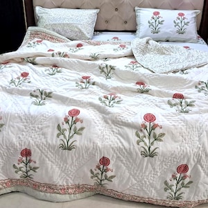 Könnte beinhalten: Weißes Blumen-Quilt-Set mit passenden Kissenbezügen. Die Bettwäsche zeigt ein sich wiederholendes Muster aus roten Blumen mit grünen Blättern. Die Steppdecke hat einen dekorativen Rand und wird auf einem Bett mit getuftetem Kopfteil präsentiert.
