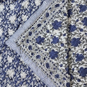 Op de afbeelding: Een blauwe en witte quilt met een bloemenpatroon met een witte achtergrond en blauwe bloemenontwerpen. De quilt heeft een rand van blauwe en witte strepen.
