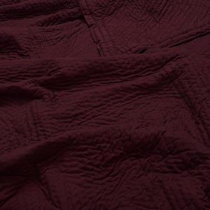 May include: A close-up shot of a burgundy-colored quilt with a textured surface. The quilt has a quilted pattern and is folded over itself, creating a layered effect. The fabric appears soft and inviting.