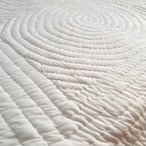 May include: A solid white quilt featuring a textured surface with intricate, repeating circular stitching patterns. The fabric displays a soft, quilted appearance with dense, concentric lines that create a tactile, rippled effect across the entire textile.