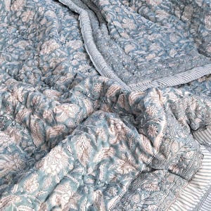 Peut inclure: Une courtepointe à motifs floraux bleus et blancs. La courtepointe présente un motif floral répétitif en blanc sur un fond bleu. Les bords sont finis avec une bordure rayée. La courtepointe est pliée et a un aspect doux et texturé.