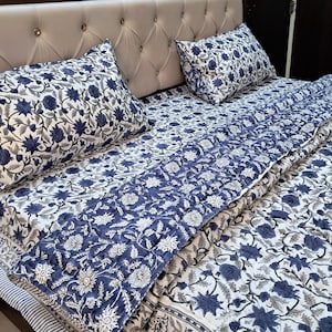 Op de afbeelding: Een bloemenbeddengoedset met een witte achtergrond en een herhaald patroon van blauwe bloemen en grijze bladeren. De set bevat twee kussenslopen en een gewatteerde deken. Het sprei heeft een blauwe rand en een gestreepte rand.