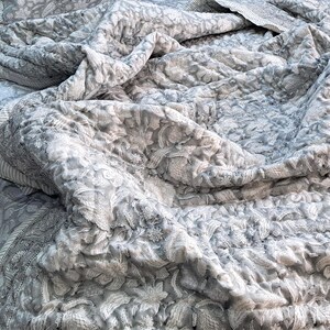 May include: A grey and white floral patterned quilt with a detailed design. The quilt has a quilted texture and a border with a striped pattern. The bedding is folded and draped, showing the intricate floral pattern.