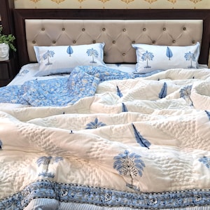 May include: A bed is made with a white and blue patterned quilt, two matching pillowcases, and a blue patterned sheet. The quilt and pillowcases feature a repeating tree and feather design. The bed is against a beige button-tufted headboard.