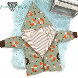 Peut inclure: Une veste à capuche verte avec une fermeture éclair et une doublure rayée marron et blanche. La veste est recouverte d'un motif de renards orange et de champignons.