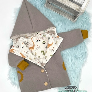 Può includere: Una giacca grigia con cappuccio e fodera con stampa di dinosauri. La giacca ha due tasche e chiusura con bottoni. Le maniche hanno polsini gialli.