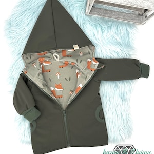 Può includere: Una giacca verde con cappuccio e chiusura a zip. L'interno della giacca è foderato con un tessuto con un motivo di volpi e funghi.