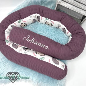 Op de afbeelding: Een paars en wit gevoerd nest met een uitneembaar kussen. Het kussen heeft de naam "Johanna" geborduurd. Het nest is versierd met een wit en groen patroon van egels.