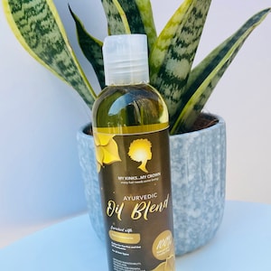 May include: A clear glass bottle with a gold label that reads "AYURVEDIC Oil Blend" and "MY KINKS...MY CROWN every hair needs some loving". The bottle is filled with a yellow liquid and is sitting on a gray surface.