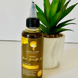 May include: A brown glass bottle with a clear dropper top, labelled "HERBAL Hair Growth Oil" with the text "MY KINKS MY CROWN every hair needs some loving" and "Enriched with Ayurvedic Herbs and Essential Oils". The bottle is sitting on a white surface next to a green potted plant.