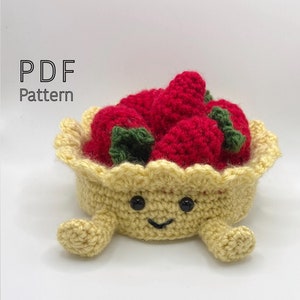 May include: A crocheted strawberry pie with a smiling face and two arms. The pie is filled with red crocheted strawberries.