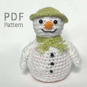 May include: A white crocheted snowman with a green hat and scarf. The snowman has black eyes and a black button nose. The text "PDF Pattern" is in the top left corner of the image.