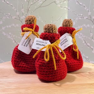 May include: Three crocheted red potion bottles with cork tops and yellow string ties. Each bottle has a white label with black text describing the potion: "Potion of Superior Healing 8d4+8", "Potion of Healing 2d4+", and "Potion of Greater Healing 4d4+4".