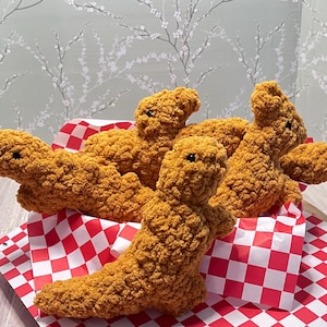May include: Four crocheted chicken nuggets in a red and white checkered paper container. The nuggets are made of a soft, fluffy yarn and are realistically shaped like chicken nuggets.