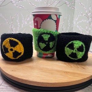 May include: Three crocheted coffee cup sleeves. Two are black with a yellow and black radiation symbol, and one is green with a green and black radiation symbol. A coffee cup is in the center, with a green sleeve.