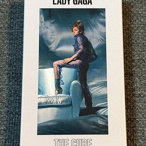 May include: A CD case with the text "LADY GAGA" at the top and "THE CURE" at the bottom. The cover features a photo of Lady Gaga in a blue outfit, posing on a light blue couch.
