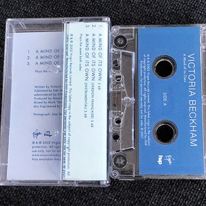 May include: A clear cassette tape case with a blue label featuring the text "VICTORIA BECKHAM" and "A Mind of its Own." The tape lists song titles and credits. The case is on a dark surface.