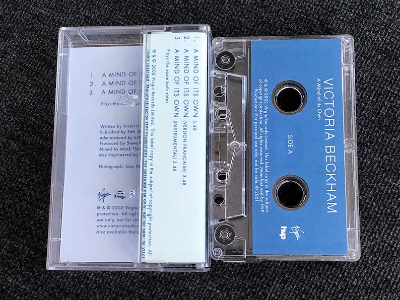 May include: A clear cassette tape case with a blue label featuring the text "VICTORIA BECKHAM" and "A Mind of its Own." The tape lists song titles and credits. The case is on a dark surface.