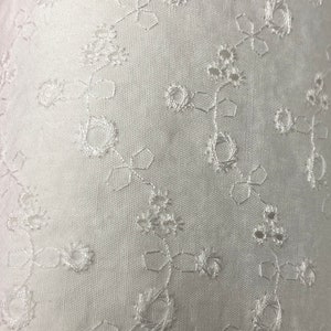 May include: White fabric with a delicate floral embroidery pattern. The fabric has a subtle texture and a soft, airy feel.