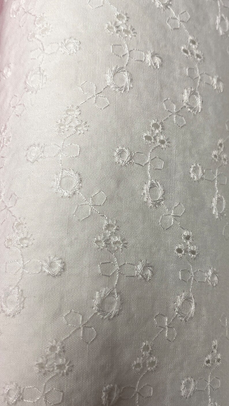 May include: White fabric with a delicate floral embroidery pattern. The fabric has a subtle texture and a soft, airy feel.