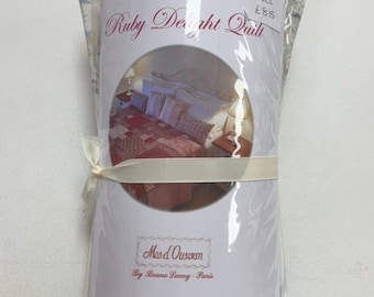 Può includere: Un kit per trapunta arrotolato intitolato Ruby Delight Quilt di Mas d'Ousvan di Bruno Lamy-Paris. La confezione presenta una fotografia circolare di un letto con una trapunta patchwork rossa e cuscini a motivi floreali blu, legata con un nastro color crema.