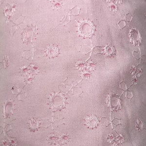 May include: A close-up of a pale pink fabric with a delicate floral pattern. The fabric has a subtle sheen and a soft, airy texture.